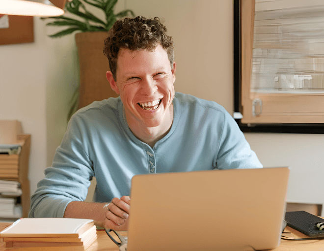A man sits at a wooden desk, eyes glued to his laptop screen, a broad smile lighting up his face in the cozy room.